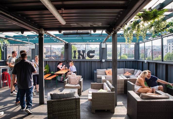 INC Sheffield - Outdoor rooftop bar with a covered seating area featuring wicker sofas with cushions, wooden tables, and string lights overhead. Green plants hang from the ceiling, and the space offers views of city buildings through mesh fencing. Several people are standing and sitting, enjoying drinks and conversation.