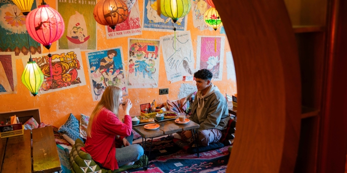 A cozy café interior with vibrant orange walls decorated with colorful Vietnamese posters and artwork. Several hanging lanterns in red, yellow, and green provide warm lighting. Two people are seated on floor cushions at a low wooden table, enjoying food and drinks. The table holds plates and cups, and the floor is covered with patterned rugs, creating a relaxed, intimate atmosphere.