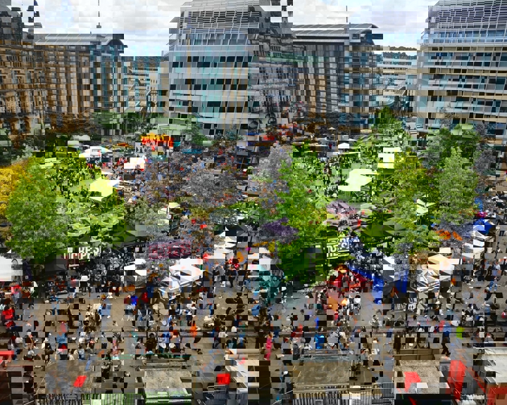 A wide aerial view of a food festival with colourful stalls, trees, and surrounding city buildings.