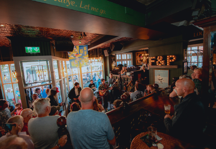 A crowded live music performance inside the Frog & Parrot pub, with warm lighting, patterned ceilings and a band playing near the front windows while people gather closely to watch.