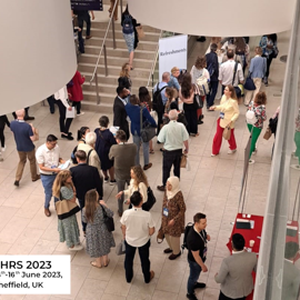 Busy conference venue with attendees networking and walking around a spacious atrium. People are gathered near a staircase and refreshment area, with tables set up for registration or materials. A sign reads “Refreshments,” and a logo in the bottom corner shows “EHRS 2023, 14th–16th June 2023, Sheffield, UK” with a white rose emblem.