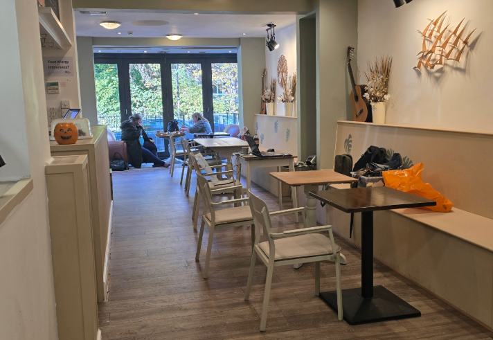 Interior of a cozy café with wooden flooring and light-colored walls decorated with dried floral arrangements and a mounted guitar. Several tables and chairs are arranged along the narrow space, with a bench running along the right wall. A small pumpkin decoration sits on a shelf to the left. Large glass doors at the back open to a view of trees outside, and a few people are seated near the windows.