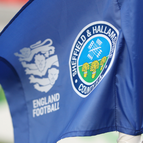 Close-up of a blue corner flag featuring the Sheffield & Hallamshire County FA crest with a white rose and crossed tools, alongside the England Football logo. The flag is positioned on a football pitch with blurred background.