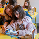 A person and a child are sat at a wooden table in a cafe, building a Lego© train together.