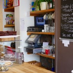 A counter in a cafe, with a glass case filled with cakes. Behind the counter is a menu written on a large blackboard.