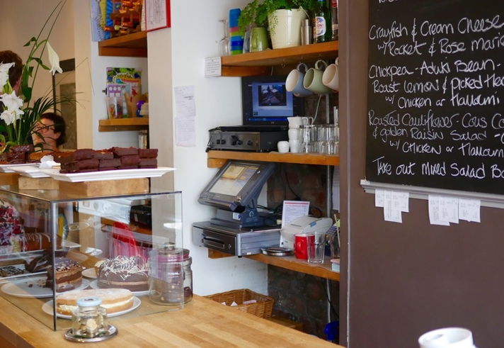 A counter in a cafe, with a glass case filled with cakes. Behind the counter is a menu written on a large blackboard.