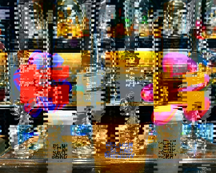 A pint of beer in a glass with the Kelham Island Brewery logo sits on a bar counter between two traditional beer pumps. The pump clips display colorful designs for two beers: one labeled “Easy Rider” Modern Blonde Ale and the other “Double Dry-Hopped Pale Ale.” The background shows shelves with liquor bottles and a warm, amber-lit backsplash.