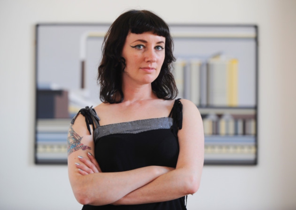 Person standing indoors with arms crossed, wearing a sleeveless black top with thin shoulder straps and a textured gray trim along the neckline. A tattoo featuring geometric and floral elements is visible on the upper arm. In the background, a framed abstract artwork with horizontal lines and muted colours hangs on a light-coloured wall.