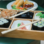 A wooden bento-style tray filled with assorted sushi, including maki rolls, nigiri topped with avocado, and sesame-coated pieces. Two pairs of chopsticks rest across the tray. In the centre, a small dish holds pickled ginger and wasabi. Behind the tray, a plate with golden spring rolls and a dipping sauce is partially visible. The food is arranged on a green table, creating a fresh and vibrant presentation.
