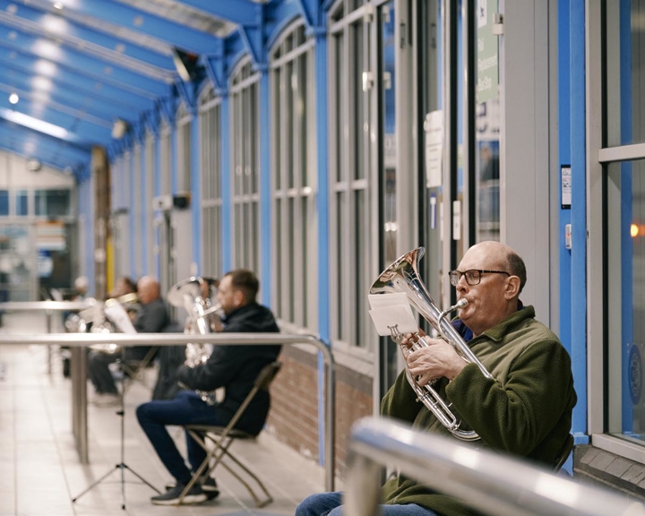 Several musicians seated in a row inside a covered walkway with bright blue metal framework and large windows, playing shiny brass instruments. The setting appears to be an indoor or semi-outdoor public space, with chairs arranged along the wall and instrument stands nearby. The scene captures a casual rehearsal or performance in a modern, well-lit environment.