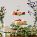 Afternoon tea at the Rhubarb Shed Cafe. On a table are a glass vase of wild flowers and a two-tier cake stand filled with cupcakes and pastries.