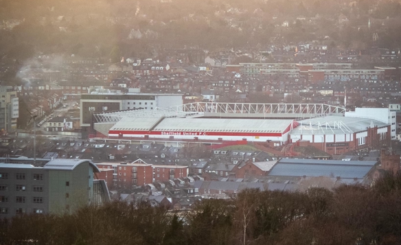 Aerial view of Bramall Lane stadium in Sheffield, surrounded by densely packed buildings and houses. The stadium has a white roof with red accents and is set against a backdrop of rolling hills under a bright, hazy sunset.