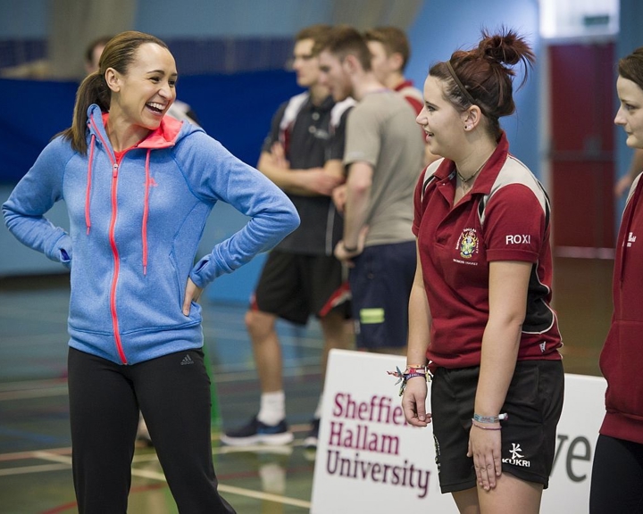 A group of people standing indoors on a sports court, wearing athletic clothing. One person in the foreground is dressed in a bright blue zip-up hoodie with pink accents and black leggings, while others wear maroon sports shirts with logos and black shorts. A white sign with red text reads “Sheffield Hallam University.” The background shows more individuals in casual sportswear and gym flooring with court markings.
