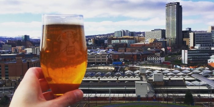 A hand holding a glass of beer in front of the Sheffield skyline on a sunny day.