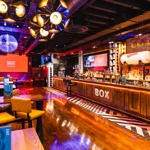 Spacious bar with long wooden counter, bold lighting, and screens displaying the venue logo