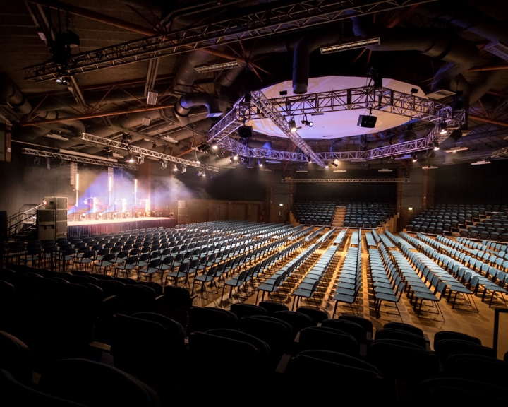 Large auditorium set up in theatre style with rows of blue chairs facing a stage. The stage is lit with spotlights and has a podium and equipment, while the ceiling features exposed beams, ducts, and a circular truss with lighting rigs. The space is dimly lit except for the illuminated stage area.