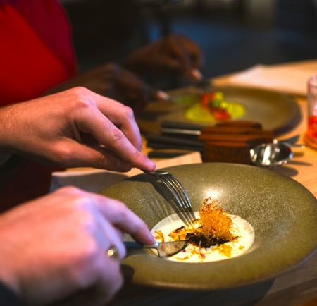 Close‑up of diners enjoying a plated fine‑dining dish at a restaurant table, with food being eaten using cutlery.