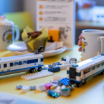 A wooden table in a cafe. On the table are two hot drinks and a partially built Lego© train, along with the instructions and the remaining pieces.