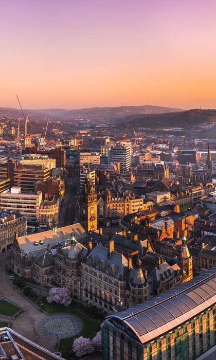 A panoramic view of Sheffield at sunrise, showcasing a mix of historic and modern architecture. The foreground features the ornate Sheffield Town Hall with its clock tower, surrounded by curved streets and green spaces. Beyond, densely packed buildings stretch across the city, with rolling hills visible in the distance under a vibrant orange and purple sky.