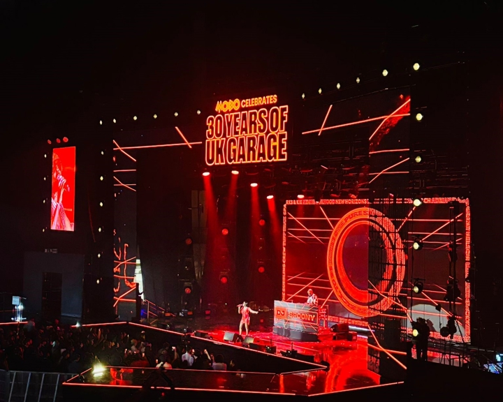 A large concert stage illuminated in red lights with geometric patterns and LED screens. At the top, bold text reads “MOBO Celebrates 30 Years of UK Garage.” A performer stands at the front of the stage, and a DJ booth labeled “DJ Spoony” is positioned behind. The audience is visible in the foreground, with spotlights and glowing wristbands creating a vibrant atmosphere inside a dark arena.