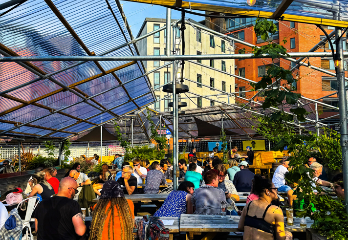  A busy outdoor seating area filled with people at wooden benches under a transparent canopy. The sun shines brightly overhead, casting strong light and highlighting surrounding greenery.