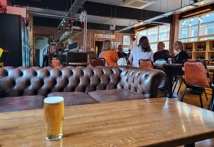 Inside at Perch Brewhouse, a pint of beer stands on a wooden table. Behind the table is a leather sofa.