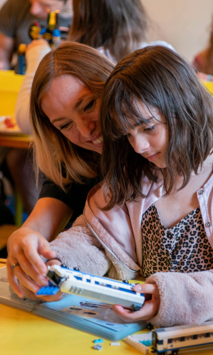 A person and a child are sat at a wooden table in a cafe, building a Lego© train together.