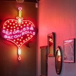 A cheeky neon sign that shows a heart pierced by a dagger with the words 'Sex, drugs & bacon rolls' on the wall at The Riverside Kelham.