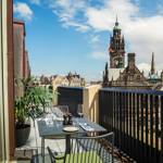 Outdoor balcony with a small dining table set for two, overlooking a historic cityscape with a tall clock tower and other ornate buildings under a partly cloudy blue sky.