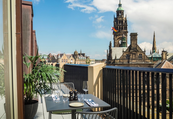 Outdoor balcony with a small dining table set for two, overlooking a historic cityscape with a tall clock tower and other ornate buildings under a partly cloudy blue sky.