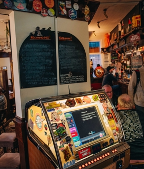 Rutland Arms jukebox lit up, with a chalk board above that's separated into forbidden and recommended. Groups of people are sat to the left of the jukebox, with the bar to the right. 